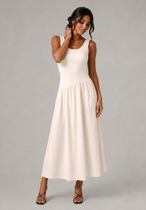 Femme portant une robe longue crème sans manches et des talons noirs à brides, debout devant un fond gris uni.