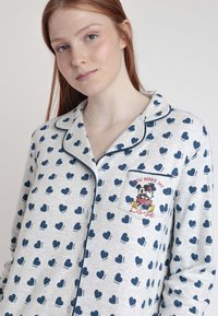 Camiseta de pijama de color gris claro con un patrón de corazones azules y ribete azul marino. Incluye un bolsillo en el pecho con un gráfico de Mickey y Minnie Mouse y el texto "TÚ HACES MI AMOR."