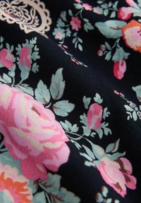 Tume kangas roosade ja korallitoonides lillemustriga, heleroheliste lehtedega ning kreemja dekoratiivse pitsdetailiga.