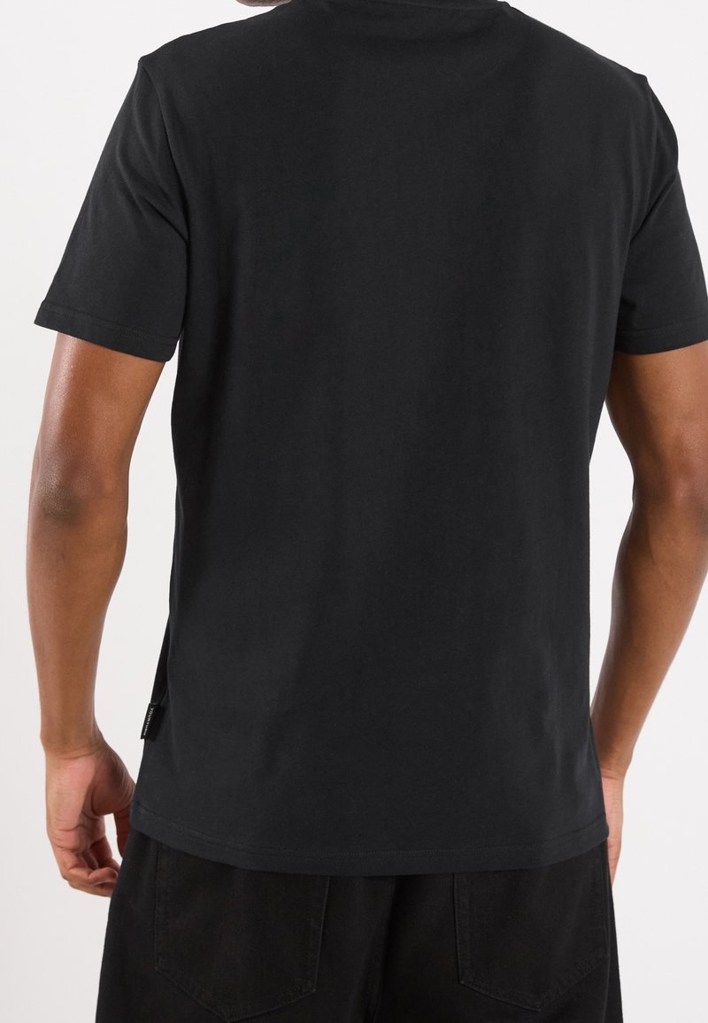 Homme portant un t-shirt noir uni à manches courtes et un pantalon noir, vu de dos contre un fond neutre.