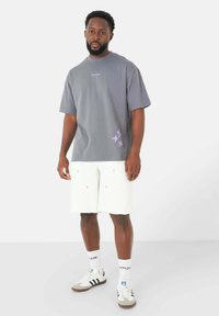 T-shirt di cotone grigio con maniche corte, caratterizzato da dettagli viola e una grafica a stelle. Abbinato a pantaloni shorts di denim bianchi e sneakers grigie.