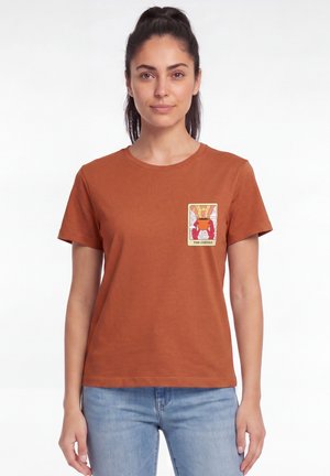 Femme aux cheveux foncés en queue de cheval portant un t-shirt couleur rouille avec un petit dessin d'une cafetière et le texte "The Coffee" sur le côté gauche de la poitrine.