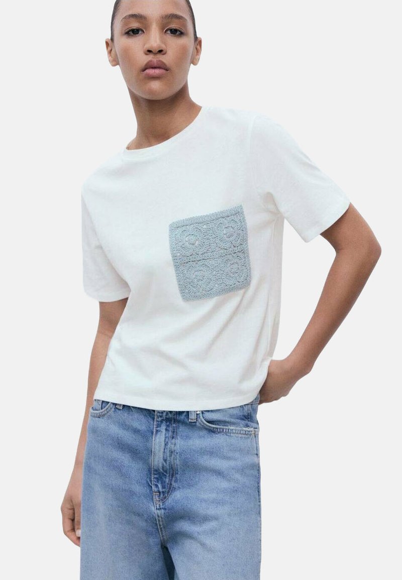 Bela bombažna majica s teksturiranim modrim kvadratnim žepom. Kratki rokavi in sproščen kroj, v kombinaciji s svetloblu denim hlačami.