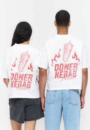 Dwie osoby noszące białe krótkie koszulki z czerwonym napisem „Döner Kebab”, grafiką obracającego się mięsa i ilustracjami płomieni na plecach.