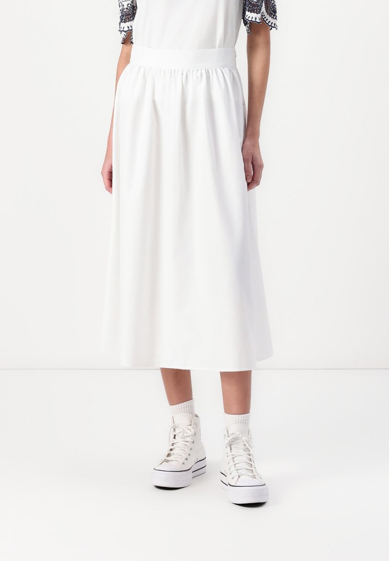 Femme portant une jupe midi blanche avec une taille élastique, associée à des baskets montantes blanches et des chaussettes blanches, sur un fond blanc uni.