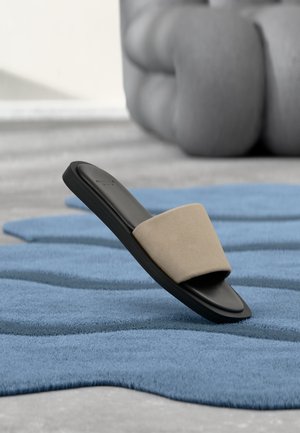 Sandale noire à glissière unique avec bride beige reposant sur un tapis bleu texturé, grand pouf gris doux en arrière-plan flou.