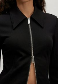 Zwarte zip-up jack met een grote kraag, metallic rits, gestructureerde stof met een gladde textuur en een getailleerd ontwerp.