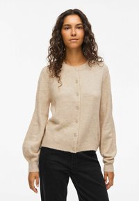 Femme aux longs cheveux bouclés portant un cardigan beige à boutons et un pantalon noir, se tenant contre un fond blanc uni.