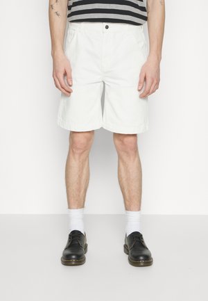 Białe bawełniane szorty o prostym kroju, z bocznymi kieszeniami i zapięciem na guzik z przodu, zestawione z czarnymi butami i białymi skarpetami za kostkę.