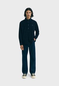 Donkerblauw overhemd met knoopsluiting, lange mouwen en een borstzak, gecombineerd met losse donkere jeans en casual schoenen met witte zolen.