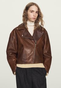 Braune Leder-Motorradjacke mit einem kurzen Design, breiten Revers, einem Reißverschluss vorne und zwei Seitentaschen. Über einem beigen Pullover getragen.