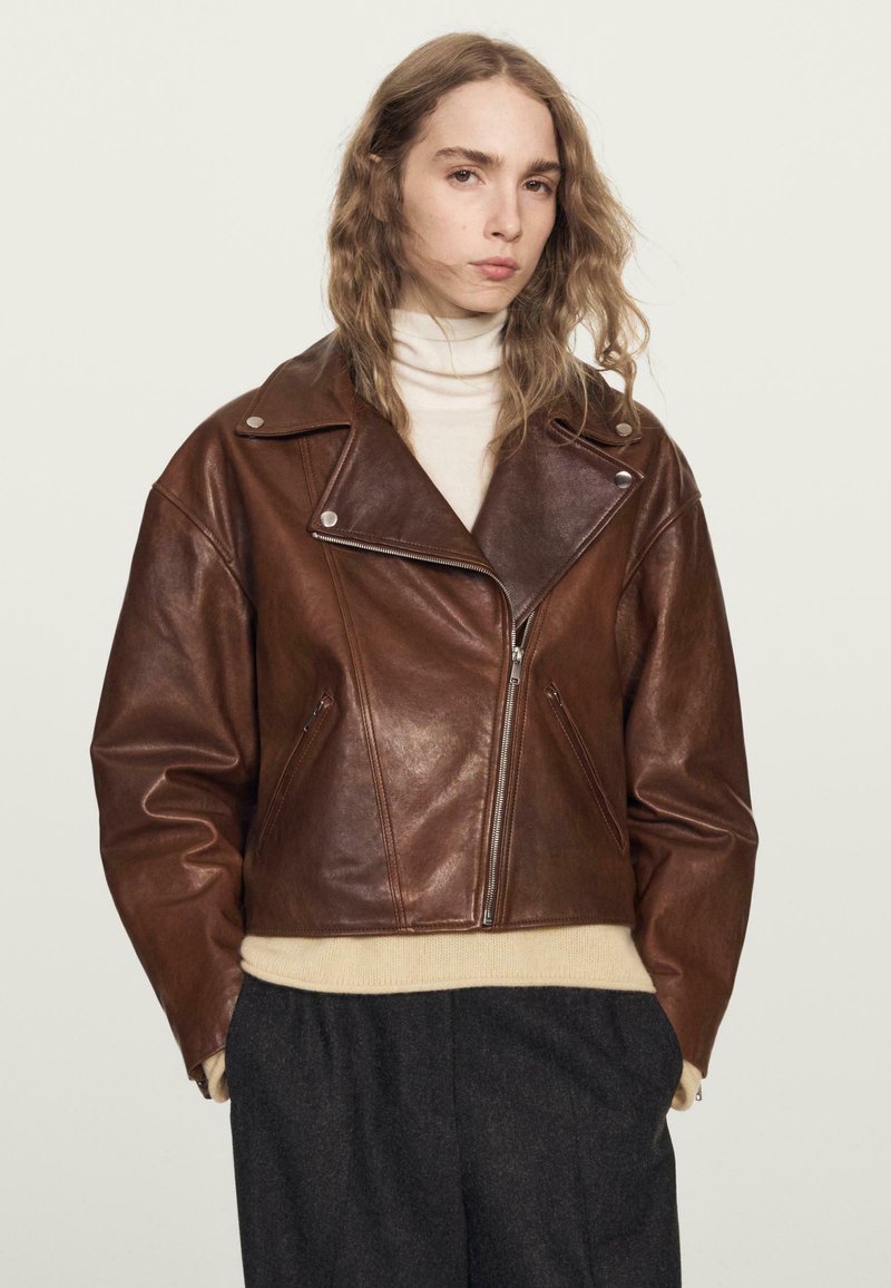 Braune Leder-Motorradjacke mit einem kurzen Design, breiten Revers, einem Reißverschluss vorne und zwei Seitentaschen. Über einem beigen Pullover getragen.