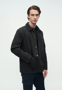 Μαύρο σακάκι από καμβά με γιακά από κορντορού, κούμπωμα μπροστά με κουμπιά, τσέπη στο στήθος και πλαϊνές τσέπες· εμφανίζεται σε μοντέλο.