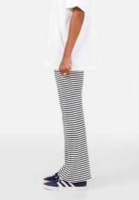 Gestreepte flared broek in marineblauw en wit, geribbelde textuur, gecombineerd met marineblauwe sneakers met witte accenten. Model draagt een wit t-shirt.