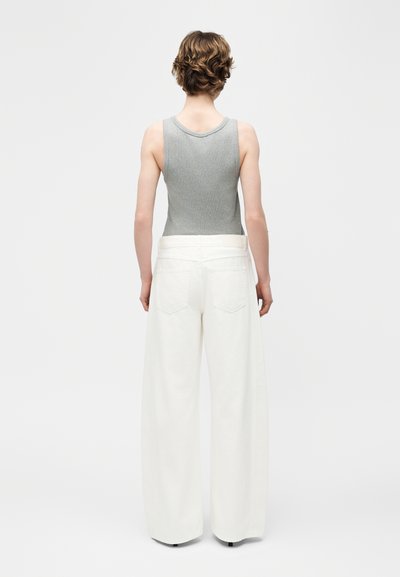 Personne aux cheveux bruns courts portant un haut gris sans manches et un pantalon blanc large, debout tournée de dos sur un fond uni.