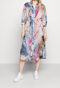 Kobieta ubrana w kolorową sukienkę midi w stylu tie-dye z wiązaniem w talii z przodu i białymi sneakersami, stojąca na tle jasnym.