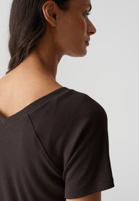 Dunkelbraunes, geripptes Oberteil mit V-Ausschnitt, kurzen Ärmeln und strukturiertem Material, das eine glatte Oberfläche an den Schultern betont.