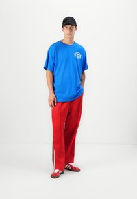 Blå bomulls-T-skjorte med logo på brystet, parret med røde treningsbukser med hvite striper. Røde joggesko fullfører antrekket.