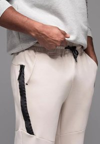 Svetle béžové tepláky s elastickým pasem a černými postranními zipy. Hladká textura, zužující se střih a viditelné detaily šití.