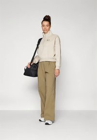 Sieviete valkā bēša krāsas pusritenti džemperi, olīvu krāsas vaļīgas plašas bikses, baltas kedas, nēsājot melnu plecu somu pret vienkrāsainu fonu.