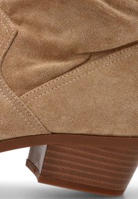 Primer plano de una bota de ante marrón claro hasta el tobillo con detalles de costura y tacón de madera apilada.
