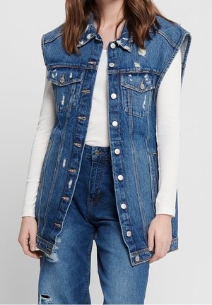 Woman wearing a sleeveless, distressed blue denim vest over a white long-sleeve top and high-waisted blue denim jeans.