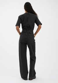 Čierny denimový overál s krátkymi rukávmi, zúženým pásom a širokými nohavicami. Obsahuje minimalistický dizajn a viditeľné švy.
