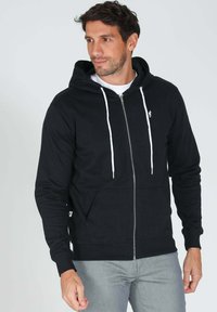 Sweat à capuche zippé noir en mélange de coton avec cordons blancs, poches avant et détail logo sur la poitrine. Coupe décontractée avec poignets côtelés.