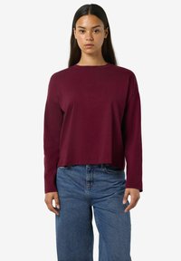 Top corto a maniche lunghe color bordeaux realizzato in tessuto morbido, con scollatura a girocollo e vestibilità rilassata, abbinato a jeans blu a vita alta.
