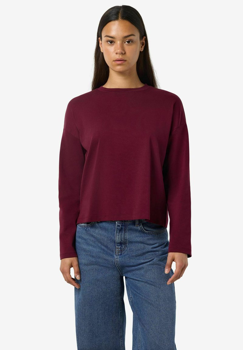 Top corto a maniche lunghe color bordeaux realizzato in tessuto morbido, con scollatura a girocollo e vestibilità rilassata, abbinato a jeans blu a vita alta.