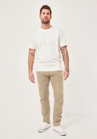 Weißes Baumwoll-T-Shirt, beige Slim-Fit-Hose, weiße Sneaker. Kurze Ärmel, runder Halsausschnitt, schlichtes Design. Tätowierungen auf einem Arm sichtbar.