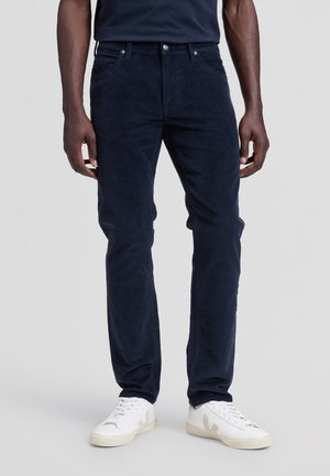 Homme portant un pantalon en velours côtelé bleu foncé et des baskets blanches, debout devant un fond uni gris clair.