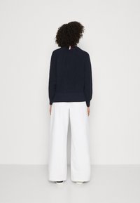Dunkelblau gestrickter Pullover mit geripptem Struktur und Raglanärmeln, kombiniert mit weißen weit geschnittenen Hosen. Akzent in Rot, Weiß und Blau am Ausschnitt.