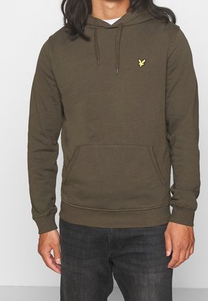 Homme portant un sweat à capuche vert olive avec poche avant et petit logo oiseau jaune sur la poitrine, associé à un jean gris foncé.