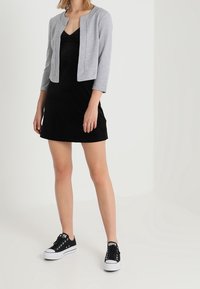 Femme portant une robe noire sans manches, une veste courte gris clair et des baskets basses noires, se tenant sur un fond neutre.