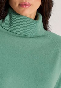 Mintgrüner Rollkragensweater aus weichem Strickstoff, mit geripptem Muster und lockerer Passform. Detail von Kragen und Nähten sichtbar.