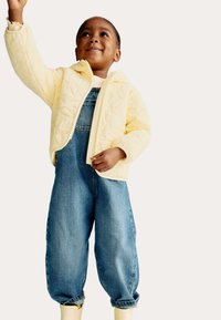 Gelbe gesteppte Jacke mit Reißverschluss, getragen über blauen Jeanslatzhosen, mit aufgeschlagenen Bündchen. Das Kind hebt einen Arm.