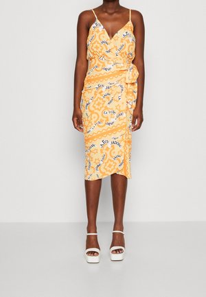 Mujer con un vestido cruzado naranja hasta la rodilla con texto con patrón blanco y negro y sandalias blancas de tacón alto contra un fondo liso.