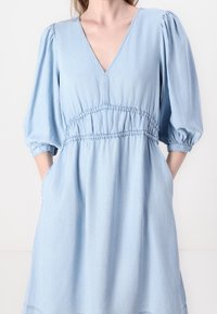Hellblaues Denim-Kleid mit V-Ausschnitt, Puffärmeln, elastischem Bund und seitlichen Taschen. Weiche Textur mit lockerer Passform und gerafften Details.