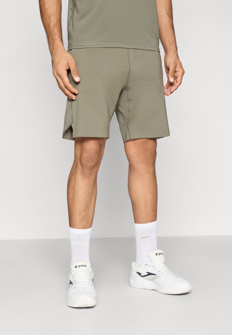 Αθλητικό σορτς σε ανοιχτό ελαιώδες πράσινο χρώμα, κατασκευασμένο από ελαστικό ύφασμα με λεία υφή και πλαϊνές σχισμές για αυξημένη κινητικότητα. Συνδυάζεται με λευκά αθλητικά παπούτσια.
