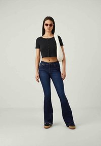 Kvinna som bär en mörk kort topp, utsvängda blå jeans, svarta platåskor, röda solglasögon och en vit axelväska, stående mot en enfärgad bakgrund.