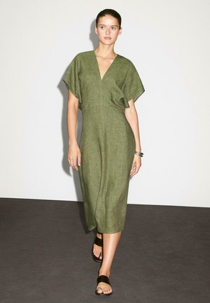 Das Modell geht selbstbewusst in einem grünen Leinenkleid und schwarzen Sandalen vor einem neutralen Hintergrund, der einen minimalistischen Modestil präsentiert.