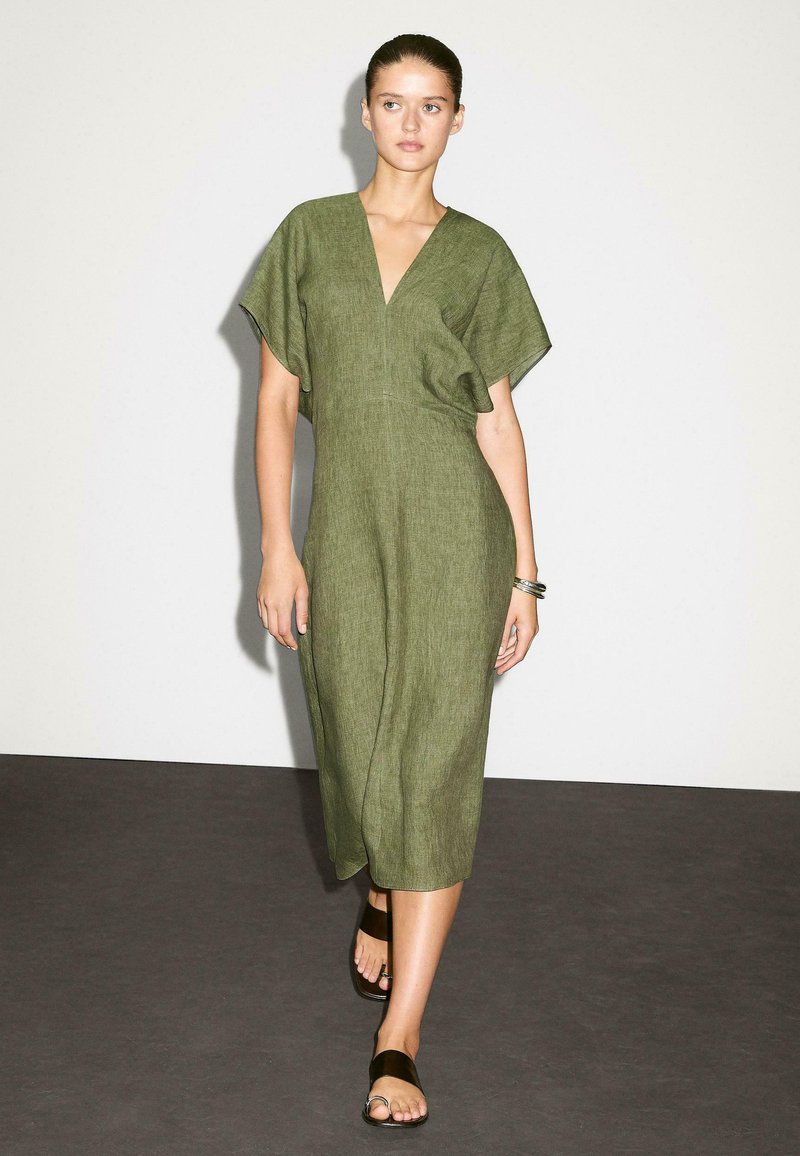 La modella cammina con sicurezza in un vestito di lino verde e sandali neri su uno sfondo neutro, mostrando un look di moda minimalista.