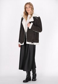 Frau mit langen blonden Haaren, die ein weißes Oberteil, eine schwarze Lammfelljacke, einen schwarzen Satinrock und schwarze Lederstiefel vor einem weißen Hintergrund trägt.