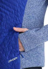 Giacca blu trapuntata con un pannello sinistro liscio e vibrante e un pannello destro testurizzato e melangiato. Presenta una tasca con zip e un logo.