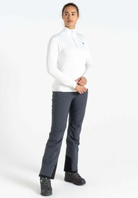 Weißes Langarmoberteil mit Reißverschlusskragen, kombiniert mit dunkelgrauen Outdoor-Hosen mit seitlichen Reißverschlüssen und strukturiertem Stoff. Schwarze Wanderschuhe.