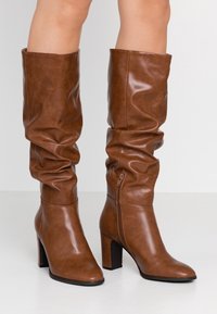 Botas de rodilla de imitación de cuero marrón con un diseño holgado, puntera cuadrada y tacón grueso. Presentan un cierre de cremallera lateral.