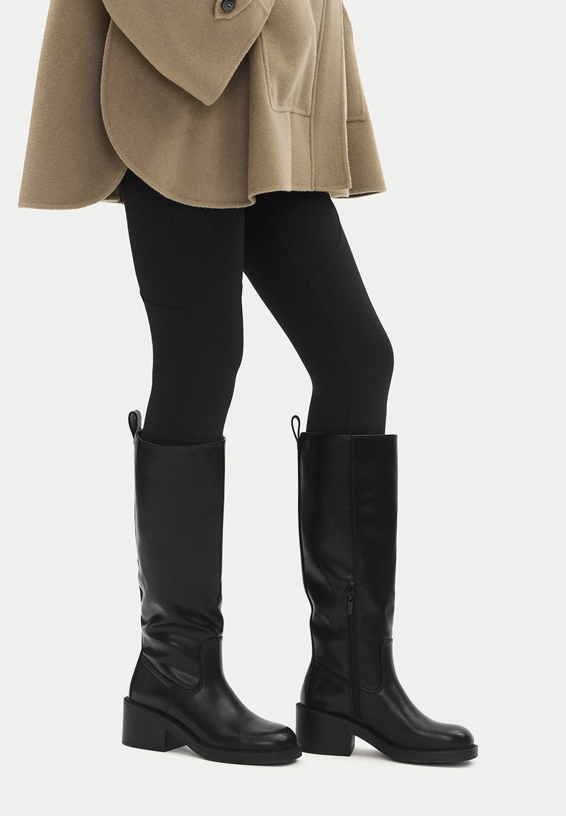 Mustad põlveni ulatuvad saapad, tehtud siledast nahast, millel on plokkkontss ja küljele paigutatud tõmblukk, kantud koos mustade retuusidega.