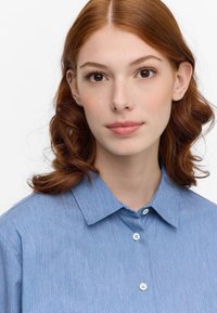 Light blue chambray shirt with a button-down collar, neat placket, and smooth texture. Features white buttons and subtle stripes in the fabric.