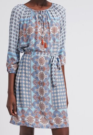 Woman wearing a light blue patterned knee-length dress with three-quarter sleeves, a drawstring neckline, and a waist tie belt.